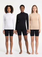 Three models standing side by side wearing long-sleeve mock neck athletic tops in white, black, and beige, paired with black biker shorts, against a plain white studio background.