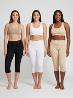 Three women standing side by side wearing matching activewear sets in three different colors: taupe, white, and beige. Each set includes a sports bra and capri leggings.