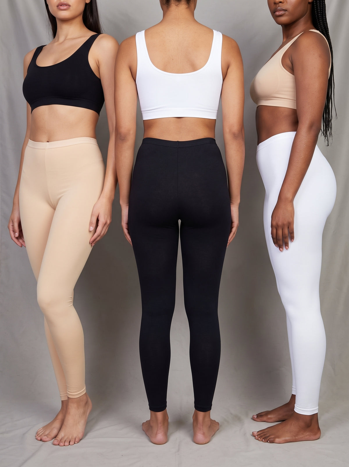 Three models standing side by side from the back wearing matching high-waisted activewear sets: a black sports bra with beige leggings, a white sports bra with black leggings, and a beige sports bra with white leggings, against a neutral gray studio backdrop.